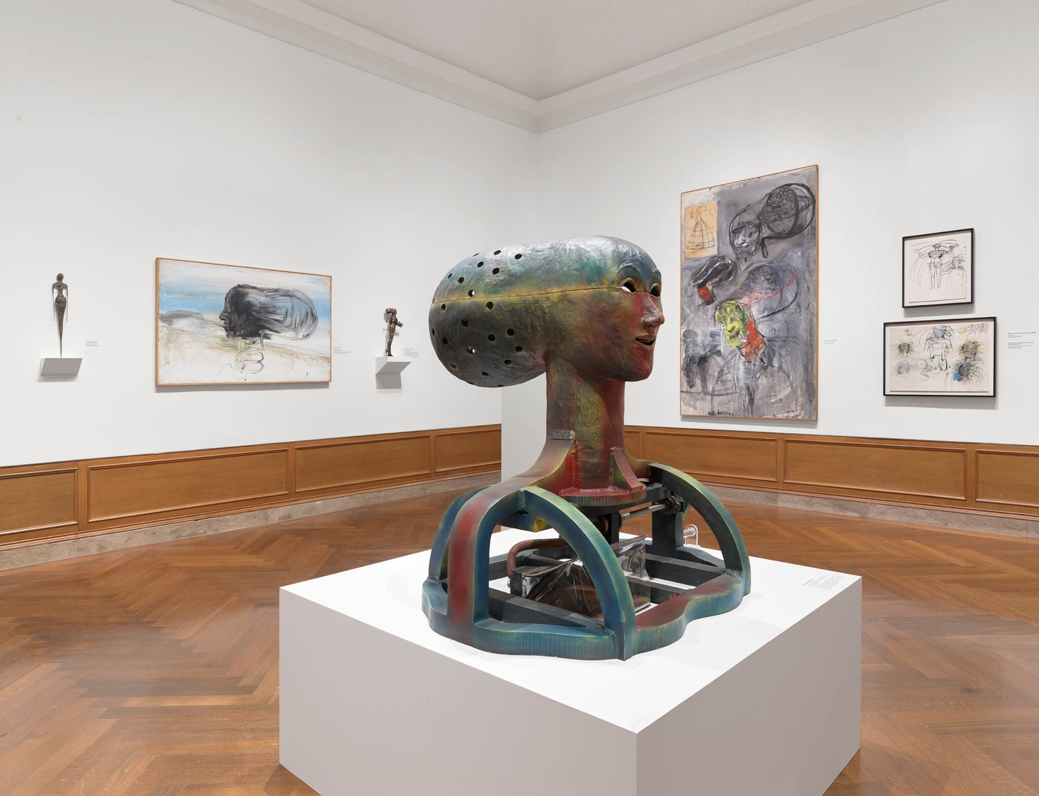 A large multi-colored head sculpture sits on a pedestal in a gallery.